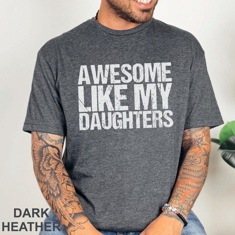 May include: Dark heather gray t-shirt with the text "AWESOME LIKE MY DAUGHTERS" in white. The shirt is a crew neck style and has short sleeves. The words "DARK HEATHER" are printed at the bottom of the image.
