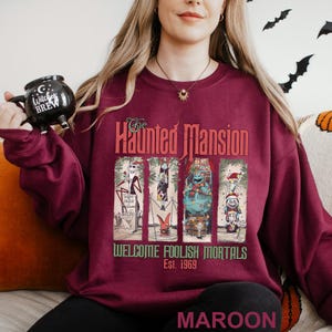 K&ouml;nnte beinhalten: Burgunderfarbenes Sweatshirt mit einem Grafikdesign von The Haunted Mansion. Das Design zeigt Charaktere und den Text "Welcome Foolish Mortals Est. 1969." Das Sweatshirt hat einen Rundhalsausschnitt.