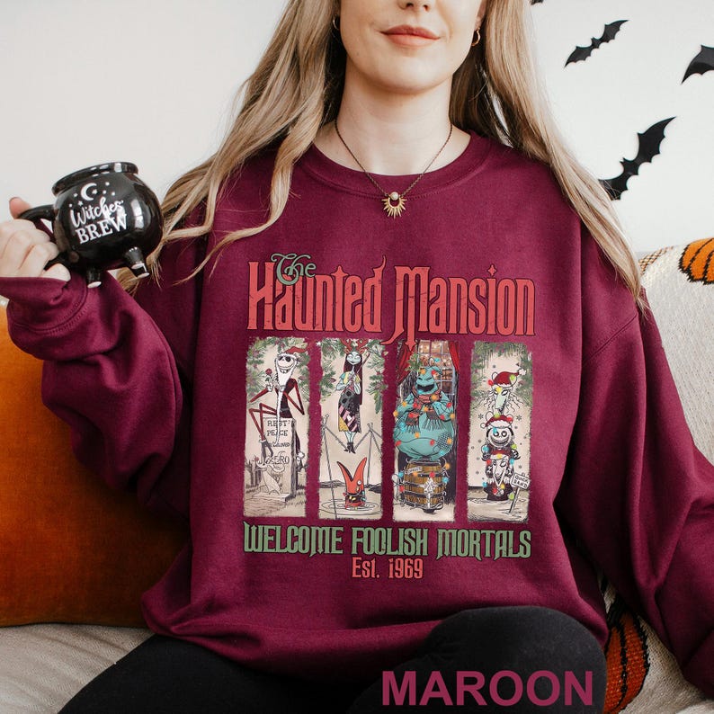 K&ouml;nnte beinhalten: Burgunderfarbenes Sweatshirt mit einem Grafikdesign von The Haunted Mansion. Das Design zeigt Charaktere und den Text "Welcome Foolish Mortals Est. 1969." Das Sweatshirt hat einen Rundhalsausschnitt.