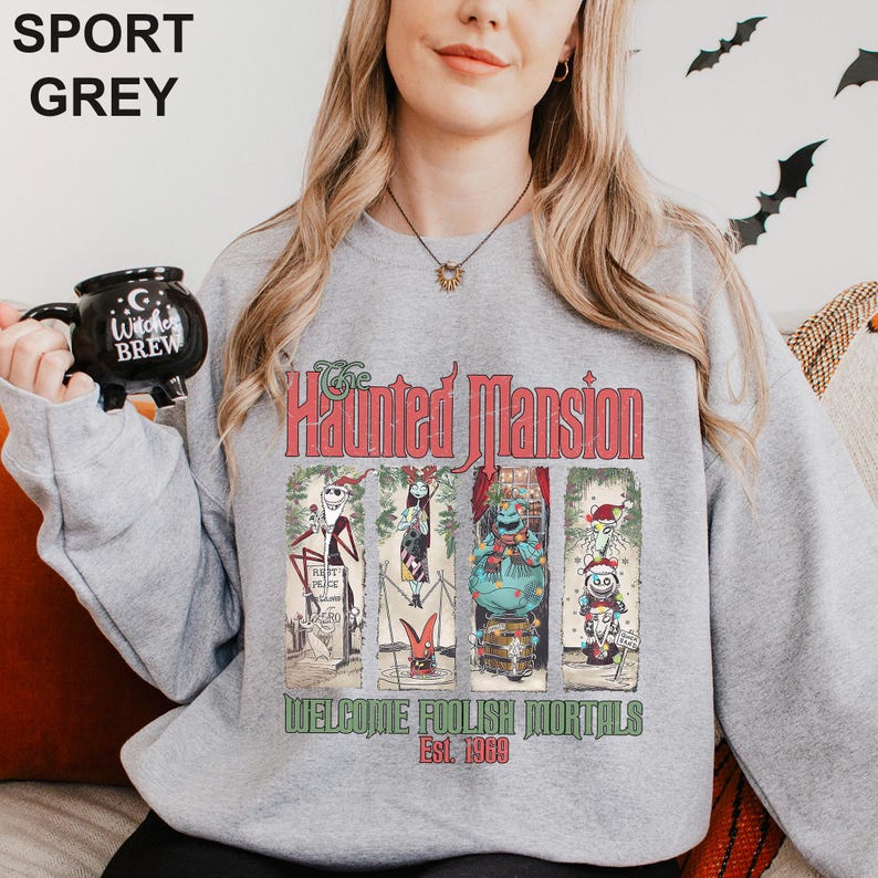 K&ouml;nnte beinhalten: Sportgraues Sweatshirt mit einem Grafikdesign von "The Haunted Mansion" und dem Text "Welcome Foolish Mortals Est. 1969". Das Design zeigt Charaktere in vertikaler Anordnung. Eine schwarze Tasse mit "Witches Brew" wird im Bild gehalten.