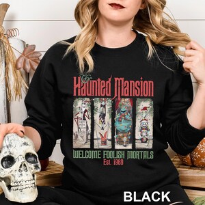 K&ouml;nnte beinhalten: Schwarzes Sweatshirt mit Rundhalsausschnitt und einem Motiv von The Haunted Mansion. Das Design enth&auml;lt den Text "The Haunted Mansion" in Rot und "Welcome Foolish Mortals Est. 1969" in Gr&uuml;n. Die Grafik zeigt vier Cartoon-Figuren.