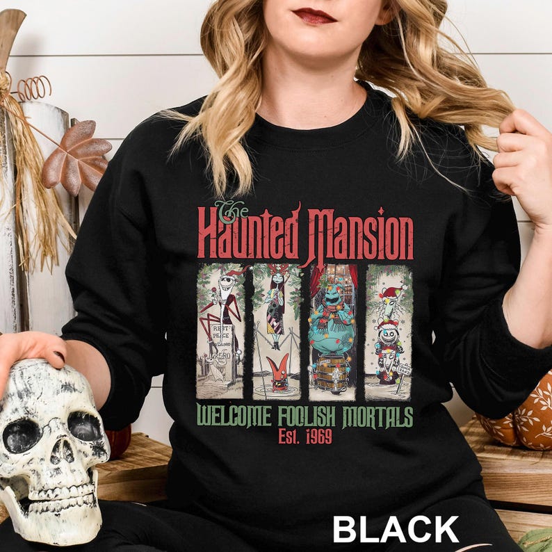 K&ouml;nnte beinhalten: Schwarzes Sweatshirt mit Rundhalsausschnitt und einem Motiv von The Haunted Mansion. Das Design enth&auml;lt den Text "The Haunted Mansion" in Rot und "Welcome Foolish Mortals Est. 1969" in Gr&uuml;n. Die Grafik zeigt vier Cartoon-Figuren.