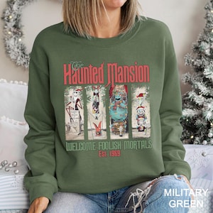 K&ouml;nnte beinhalten: Milit&auml;rgr&uuml;nes Sweatshirt mit einem Grafikdesign von "The Haunted Mansion". Das Design enth&auml;lt den Text "The Haunted Mansion" in Rot und "Welcome Foolish Mortals Est. 1969". Die Grafik zeigt Cartoon-Charaktere in einem vertikalen Panel-Design.