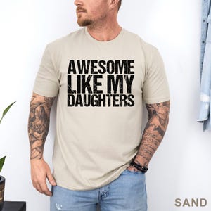 May include: Sand-colored t-shirt with the text "AWESOME LIKE MY DAUGHTERS" in bold, black letters. The shirt has short sleeves and a crew neck. The word "SAND" is printed in the bottom right corner.