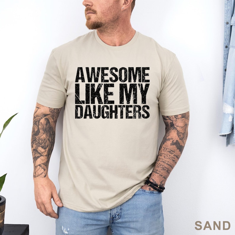 May include: Sand-colored t-shirt with the text "AWESOME LIKE MY DAUGHTERS" in bold, black letters. The shirt has short sleeves and a crew neck. The word "SAND" is printed in the bottom right corner.
