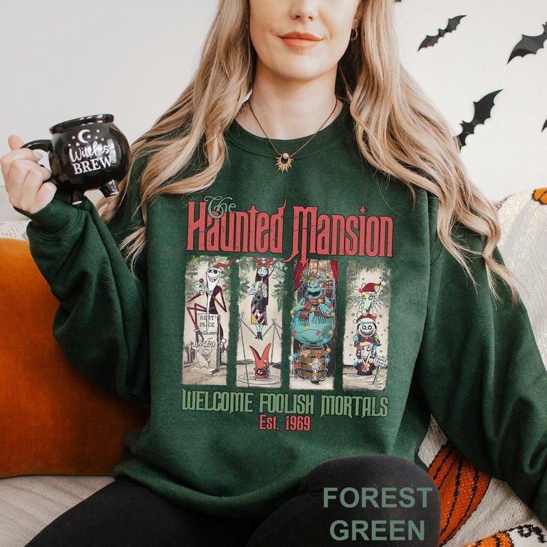 K&ouml;nnte beinhalten: Forstgr&uuml;nes Sweatshirt mit einem Haunted Mansion-Motiv. Das Design enth&auml;lt die Worte "Haunted Mansion" und "Welcome Foolish Mortals Est. 1969" mit Cartoon-Charakteren. Das Sweatshirt hat eine einfarbige forstgr&uuml;ne Farbe.