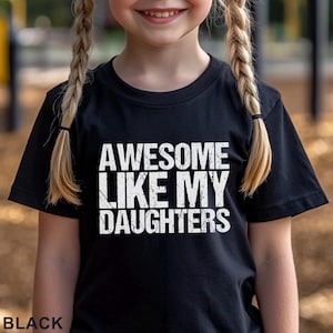 May include: Black t-shirt with the text "AWESOME LIKE MY DAUGHTERS" in white, distressed lettering. The word "BLACK" is printed at the bottom of the shirt. The shirt is being worn by a person with blonde braids.