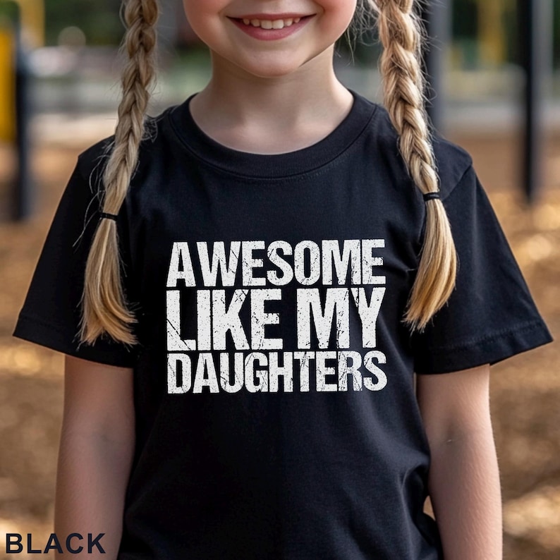 May include: Black t-shirt with the text "AWESOME LIKE MY DAUGHTERS" in white, distressed lettering. The word "BLACK" is printed at the bottom of the shirt. The shirt is being worn by a person with blonde braids.