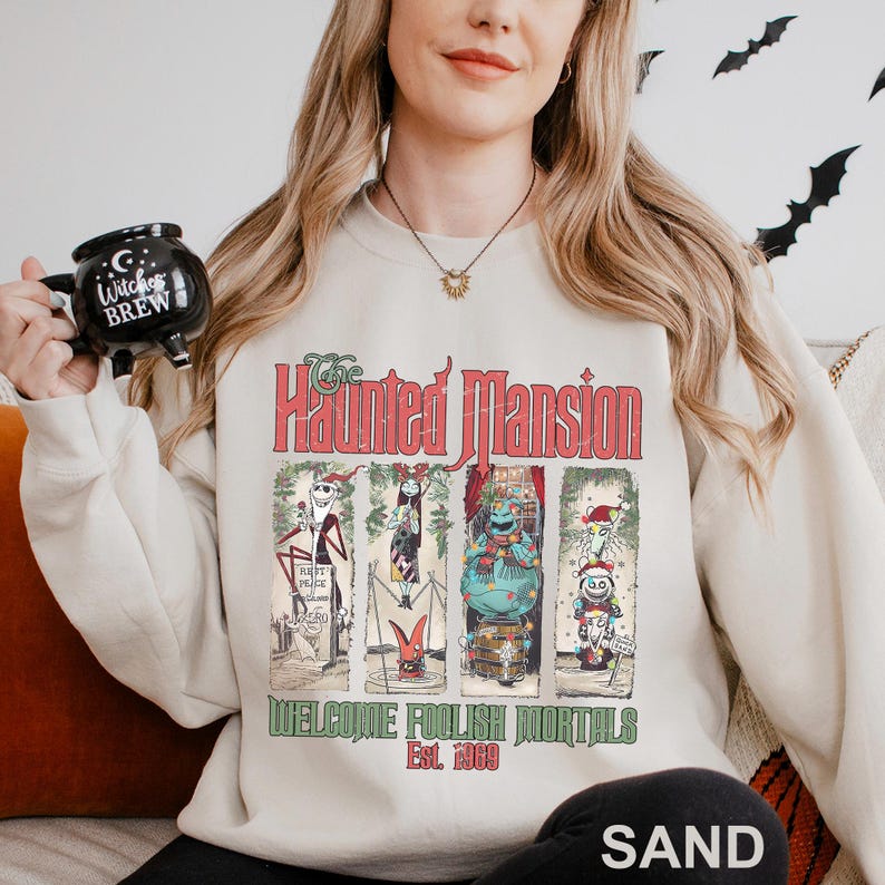 K&ouml;nnte beinhalten: Sandfarbenes Sweatshirt mit einem Grafikdesign der Haunted Mansion. Das Design enth&auml;lt die roten Worte "Haunted Mansion" und "Welcome Foolish Mortals Est. 1969". Die Grafik zeigt vier gerahmte Bilder von Charakteren.