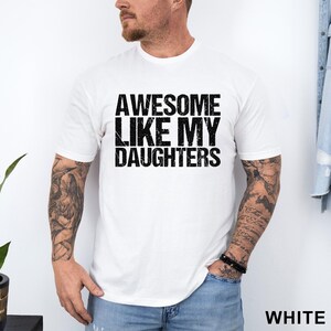May include: White t-shirt with the text "AWESOME LIKE MY DAUGHTERS" in a distressed black font. The shirt is short-sleeved and has a crew neck. The word "WHITE" is printed in the bottom right corner.