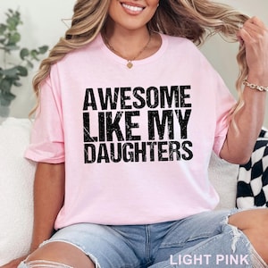 May include: Light pink t-shirt with the text "AWESOME LIKE MY DAUGHTERS" in black. The shirt is a crew neck style and is made of a soft material. The words "LIGHT PINK" are printed at the bottom of the image.