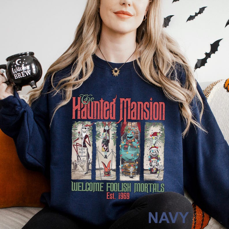 K&ouml;nnte beinhalten: Marineblaues Sweatshirt mit einem Grafikdesign von The Haunted Mansion. Das Design enth&auml;lt Charaktere und den Text "WELCOME FOOLISH MORTALS Est. 1969". Ein bequemer, l&auml;ssiger Stil.