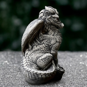 Peut inclure: Une petite sculpture de dragon en pierre grise avec des ailes repliées derrière son dos. Le dragon est assis sur une base de roche et a une surface texturée détaillée.