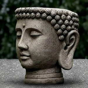 May include: A grey stone Buddha head planter with closed eyes and detailed curly hair. The sculpture is a serene representation of Buddha, suitable for indoor or outdoor decor. The planter is made of stone.