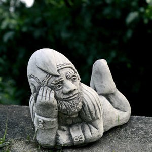 May include: A gray concrete garden gnome statue in a relaxed pose, resting on its elbow. The gnome has a long beard, a pointed hat, and is wearing a tunic. The statue is set against a blurred green background.