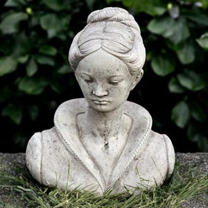 May include: A weathered stone bust of a woman with her head bowed. The sculpture features detailed hair and a high collar. The bust is set against a backdrop of green foliage and rests on a stone surface with grass.