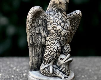 May include: A detailed, gray stone eagle statue with outstretched wings, clutching a fish. The sculpture has intricate feather detailing and stands on a round base. The eagle's beak is slightly open, and its eyes are visible.