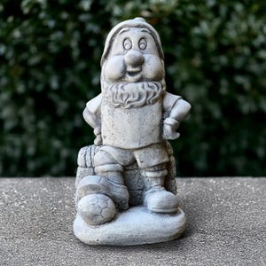 Puede incluir: Una figura de gnomo de jardín de hormigón gris que lleva una camiseta y pantalones cortos, sosteniendo un balón de fútbol y de pie sobre una base.