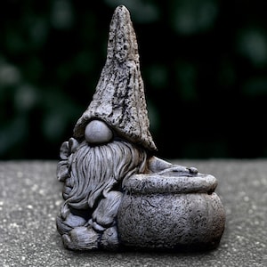 Può includere: Statua di gnomo da giardino in cemento grigio con un lungo cappello a punta e una lunga barba fluente. Lo gnomo tiene in mano un vaso strutturato. La statua ha un aspetto ruvido e invecchiato, che suggerisce che sia fatta di pietra.