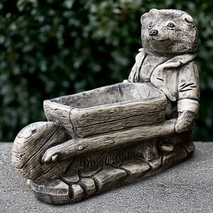 May include: A detailed, gray-toned garden planter shaped like a bear pushing a wheelbarrow. The bear wears a coat and the wheelbarrow has a wooden texture. The planter is made of a durable material, suitable for outdoor use.