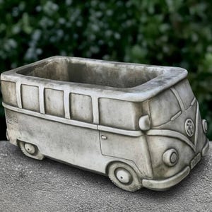 May include: A gray, weathered concrete planter shaped like a vintage camper van. The rectangular vessel features molded details including circular headlights, a front emblem, side windows, and wheels, with a hollow interior for planting.