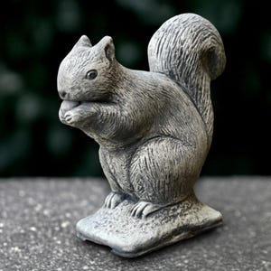 Puede incluir: Estatua de ardilla de piedra gris, sosteniendo una nuez en sus patas. La ardilla tiene una cola tupida y está sentada sobre una pequeña base texturizada. Estatua detallada y realista, adecuada para la decoración del jardín.