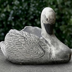 Könnte beinhalten: Ein verwitterter, grauer Schwan-Pflanzkübel mit detaillierten Feder- und Flügelmustern. Der Schwan hat einen schwarzen Schnabel und eine ausgehöhlte Rückseite zum Bepflanzen. Die Skulptur besteht aus einem steinartigen Material und ist für den Außenbereich geeignet.
