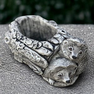 May include: A gray, stone-like planter shaped like a log, with two hedgehogs sculpted on the side. The planter has a hollowed-out center for plants and is decorated with leaf and vine details. The hedgehogs have detailed fur and expressive eyes.