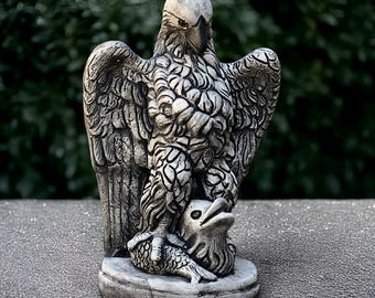 May include: A detailed, gray-toned statue of an eagle with outstretched wings, perched atop a fish and bird. The sculpture has intricate feather detailing and stands on a circular base. The background is blurred green foliage.