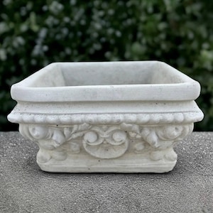 Può includere: Un vaso quadrato color bianco sporco con un bordo largo e piatto e una base decorativa caratterizzata da volute intagliate e motivi floreali in rilievo. Il vaso presenta una finitura opaca liscia e un interno profondo e aperto dal design classico.