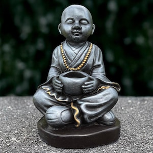 May include: A small, grey, meditating Buddha statue with a gold beaded necklace and a bowl in its hands. The statue is sitting on a black base.
