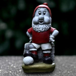 Puede incluir: Figura de gnomo de jardín de piedra, con camiseta roja, pantalones cortos blancos y zapatos negros, con barba blanca y gorro rojo. El gnomo está junto a un balón de fútbol. La figura tiene una expresión sonriente.