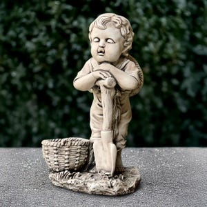 May include: A beige garden statue of a young child leaning on a shovel, with a woven basket at its feet. The child is wearing a hat and overalls. The statue is on a stone base, with a blurred green background.