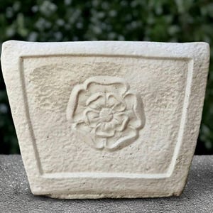 May include: A square, off-white planter with a textured surface. The front features a raised floral design. The planter is made of a durable material and is suitable for indoor or outdoor use.