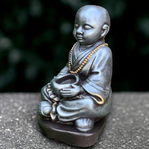 May include: A small, seated statue of a young monk in a meditative pose. The figure is dark grey with gold accents on the necklace and robe. The monk holds a small bowl and sits on a dark base.