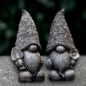 Puede incluir: Dos gnomos de jardín grises con sombreros texturizados y largas barbas. Un gnomo sostiene una pequeña pala, el otro una flor. Los gnomos están sobre una superficie gris con un fondo verde borroso.