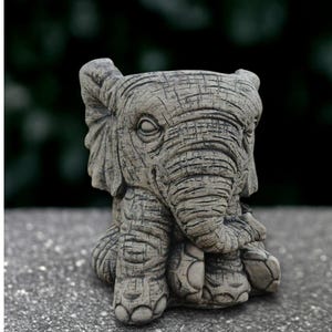 May include: A gray elephant-shaped planter with a textured surface. The elephant is sitting with its legs tucked under its body. The planter has a hollowed-out top for holding plants or other items. The elephant's features include large ears, eyes, and a trunk.