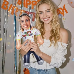 May include: A woman holding a personalized birthday balloon in the shape of a person. The balloon has a white shirt with colorful sprinkles and the words "Happy Birthday." The balloon has blue jeans and silver shoes. The background includes a birthday banner.