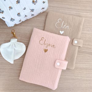 May include: A selection of baby items. A pink and beige fabric journal with gold embroidered names, a white floral cosmetic bag, and a white heart-shaped keychain are visible.