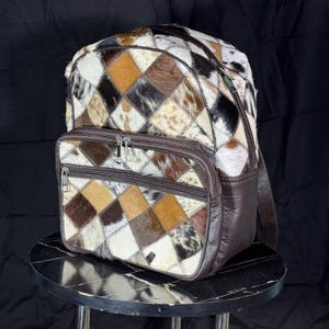 Può includere: Uno zaino patchwork con una tasca frontale con cerniera. La borsa presenta un motivo a rombi di pelle di animale marrone, beige, nero e bianco. I lati e le cinghie sono in pelle marrone scuro. Lo zaino è appoggiato su un tavolo di marmo bianco e nero.