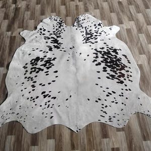 May include: A cowhide rug featuring a natural, irregular shape with a white base color and scattered black spots of varying sizes across the surface. The hide displays a smooth texture with a distinct pattern of dark markings throughout the center.