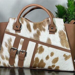 May include: A brown and white cowhide handbag with a brown leather trim and double buckles. The bag has two top handles and a detachable shoulder strap.