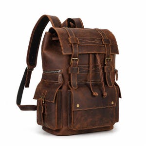 May include: A dark brown leather backpack featuring a top flap with two buckle straps, a drawstring closure, and multiple exterior pockets with metal hardware. The bag includes adjustable shoulder straps and visible stitching details throughout.