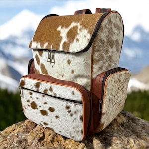 Op de afbeelding: Een bruine en witte koeienhuid rugzak met bruine lederen accenten. De rugzak heeft een groot hoofdvak en twee kleinere zakken aan de voorkant.