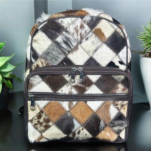 May include: A patchwork backpack and matching pouch set. The bags feature a diamond pattern made of various shades of brown, white, and black fur. The backpack has a top handle and adjustable straps. The pouch has a zipper closure.