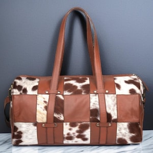 May include: A brown leather and cowhide patchwork tote bag with two top handles. The bag has a checkered pattern with brown leather squares and white and brown cowhide squares.