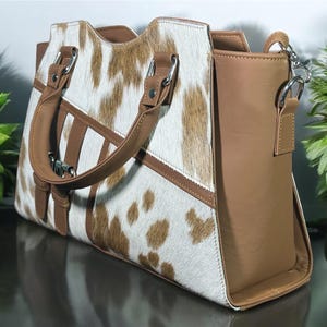 May include: A brown and white cowhide handbag with a brown leather strap and handle. The bag has a brown leather flap with a silver buckle.