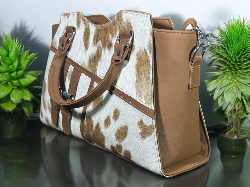May include: A brown and white cowhide handbag with a brown leather strap and handle. The bag has a brown leather flap with a silver buckle.