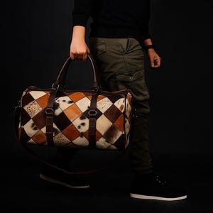 May include: A brown and tan leather duffel bag with a patchwork design. The bag features a diamond pattern with various shades of brown, tan, and white. It has brown leather handles and a shoulder strap. The bag is being carried.