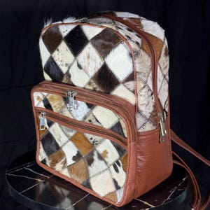 May include: A patchwork backpack with a brown leather base and straps. The upper portion features a diamond pattern of brown, black, and white animal hide. The bag has multiple zippered compartments and silver-toned hardware.
